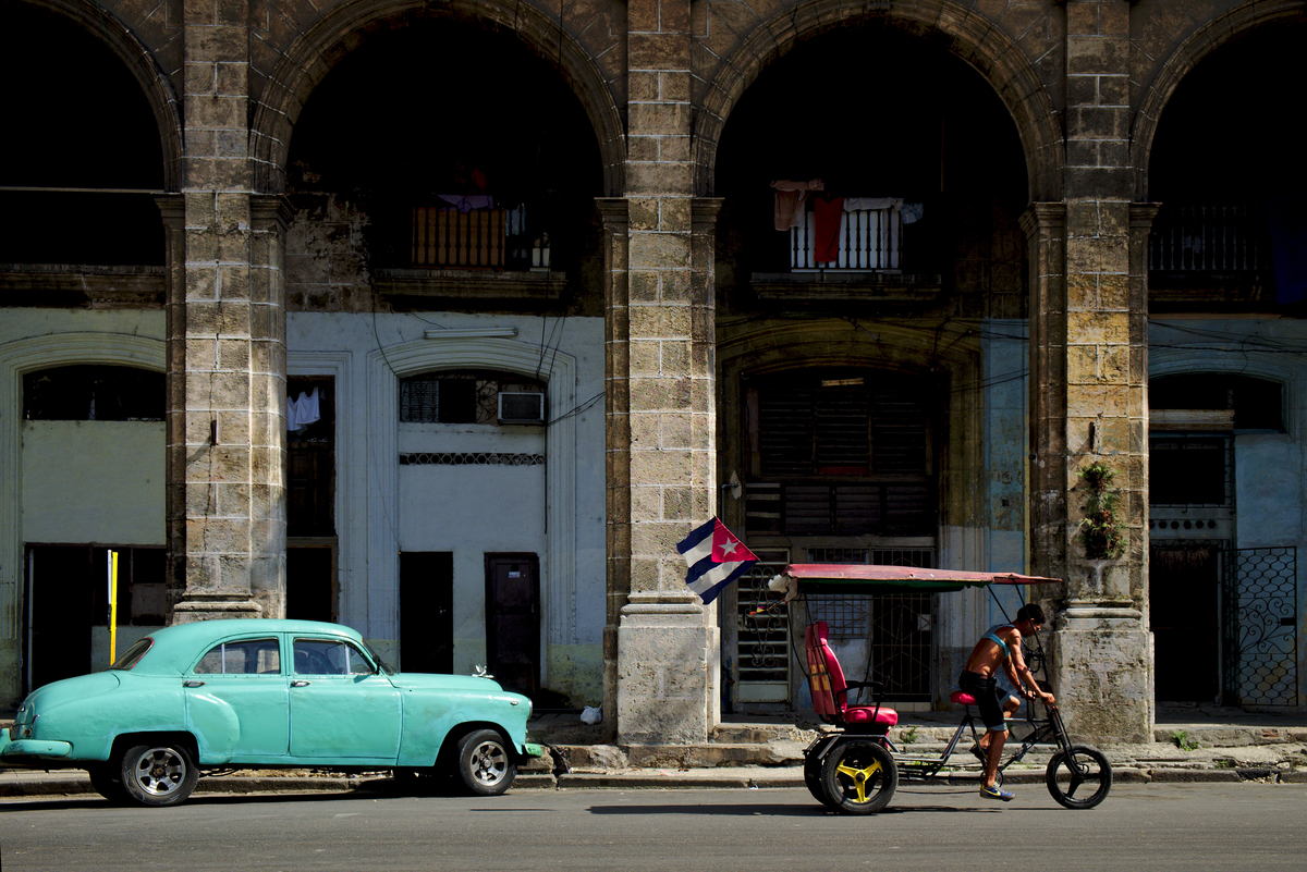 Um velho carro americano estacionado nas grandes arcadas típicas de Centro Habana, enquanto passa um bicitaxi.